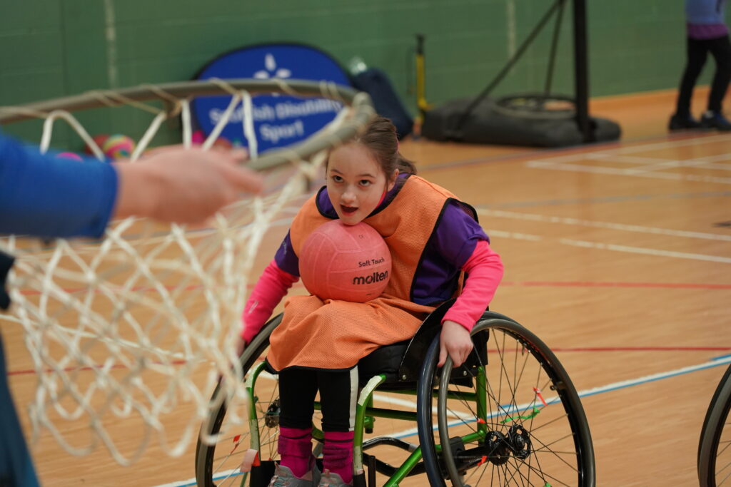 Young girl in a wheelchair playing basketball. She is balancing the basketball on her lap as she gets into a good position to shoot for the hoop.