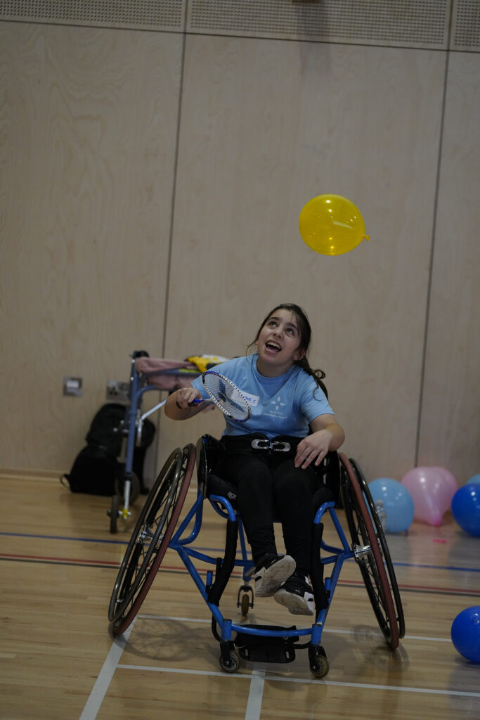 Photo of a young girl in a wheelchair playing badminton with a racquet and yellow balloon.